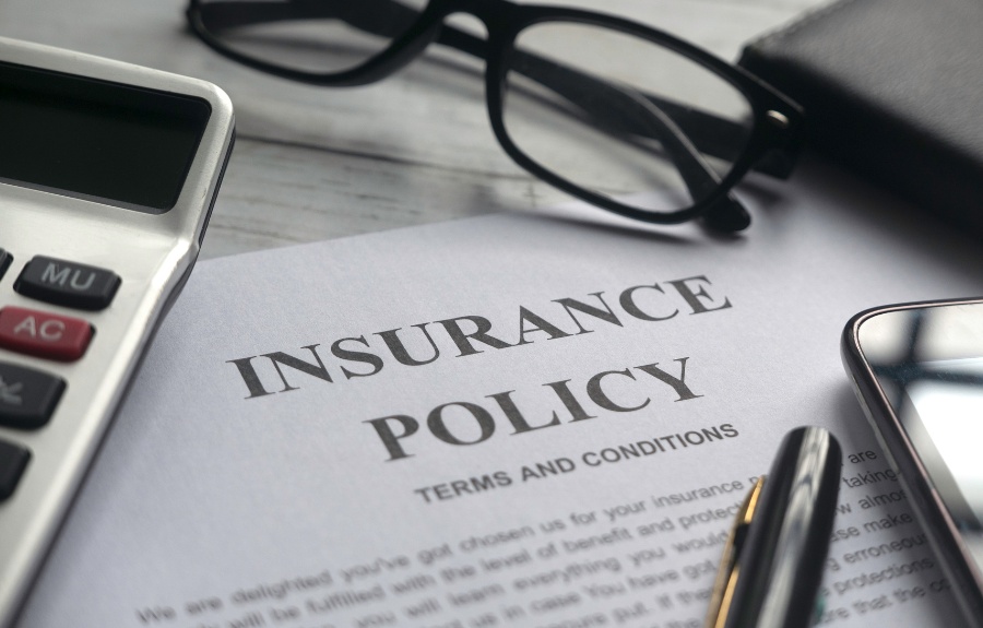 Close-up of an insurance policy document with terms and conditions visible, next to reading glasses, a calculator, pen, and smartphone on a desk.