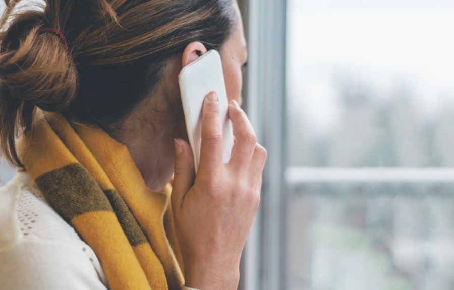 Woman in a cozy yellow plaid scarf makes a phone call while looking out a window on a gray winter day.